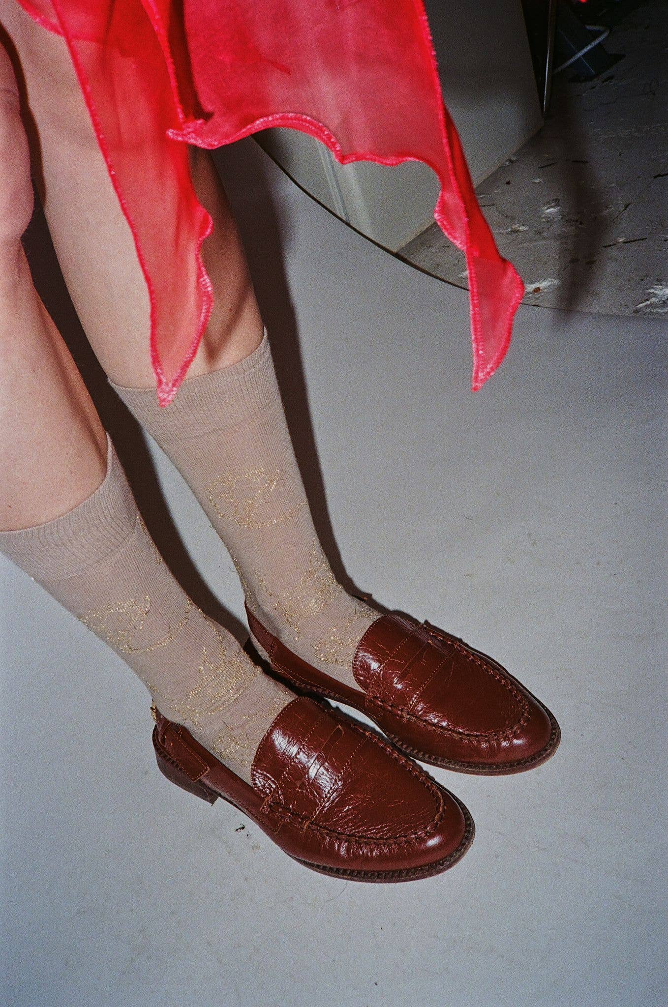 Brown loafers worn with beige socks and a red garment on a plain background