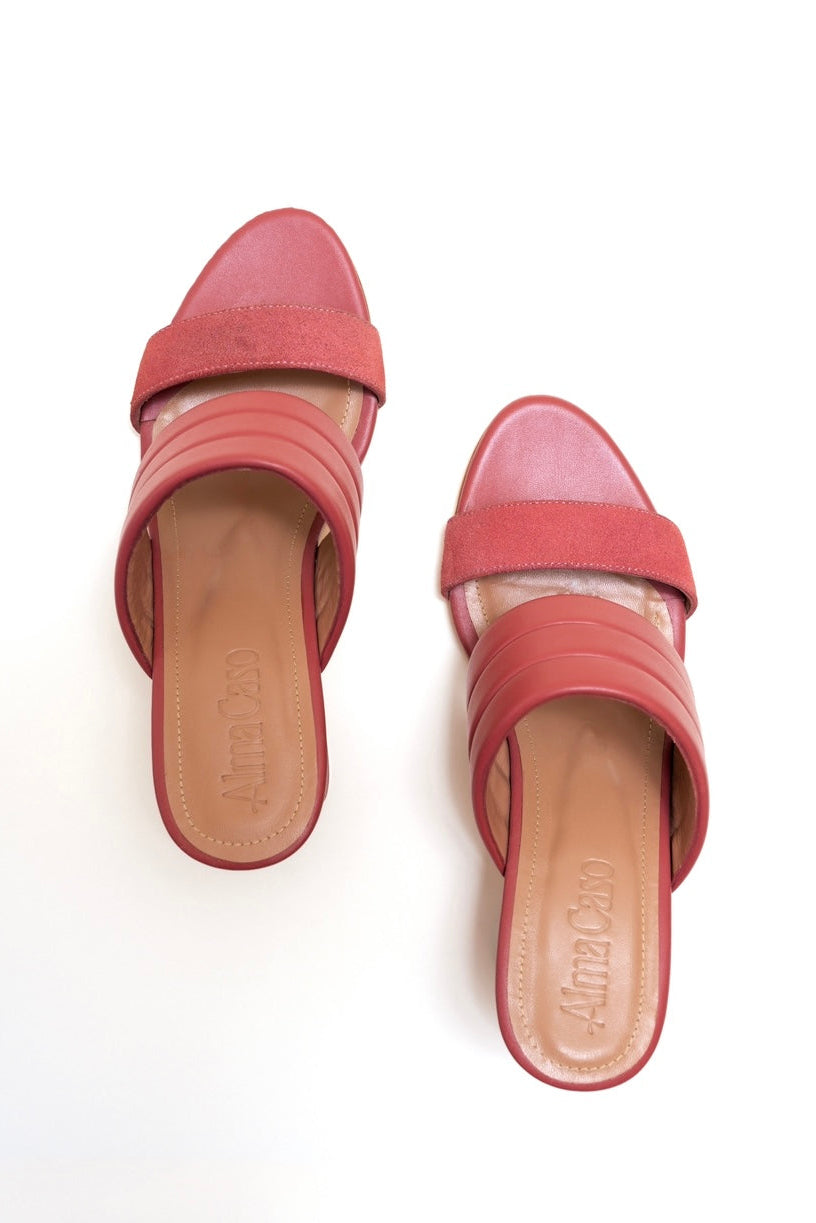 A pair of coral pink slide sandals with wide double straps and low block heels, shown from a top-down view on a white background. The brand name 'Ami Cosé' is embossed on the tan insoles.