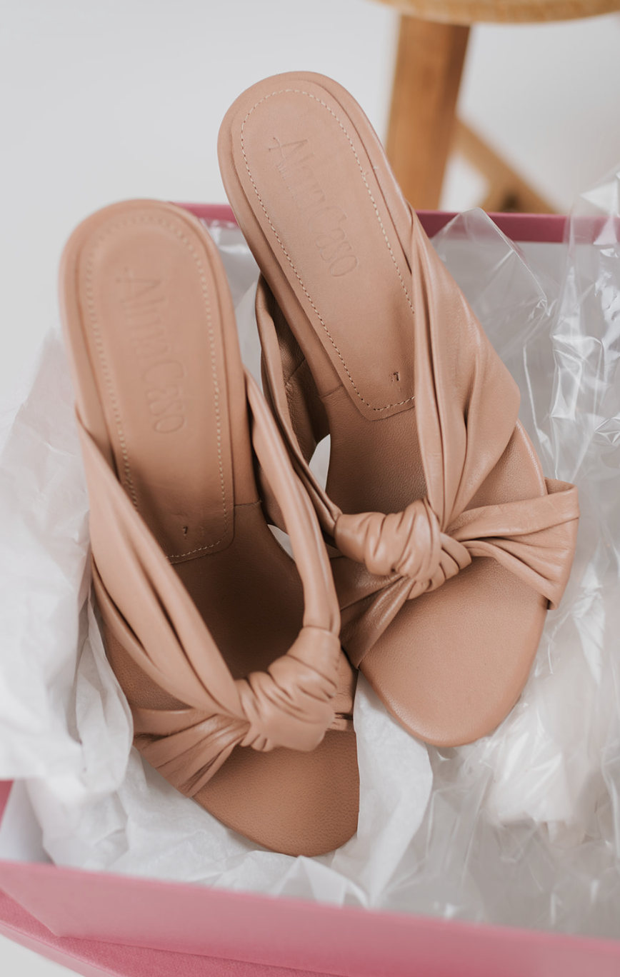 A pair of nude beige knotted leather mule sandals with knotted front straps, resting in a pink shoebox lined with white tissue paper.