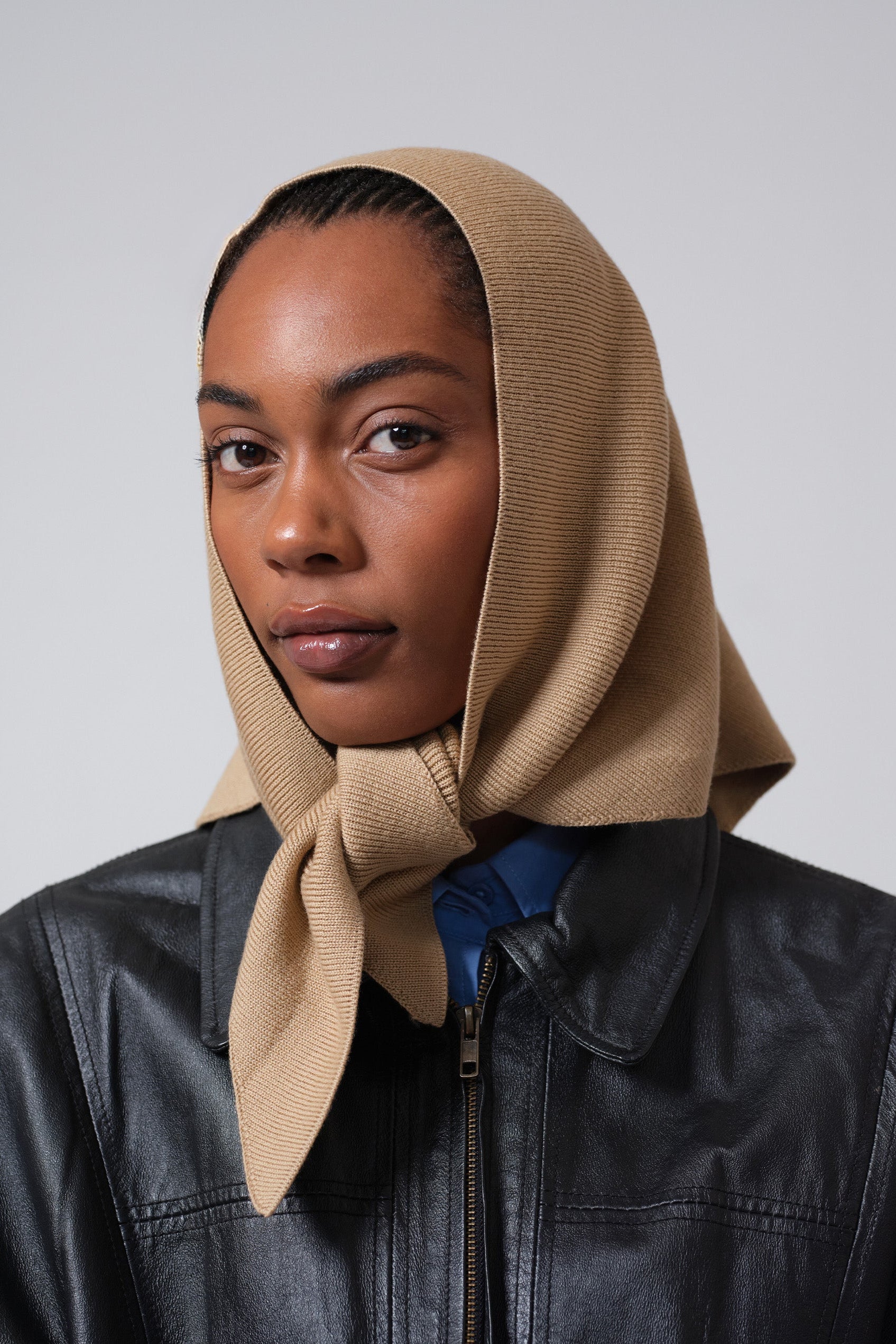 Woman wearing a beige headscarf and black jacket against a gray background