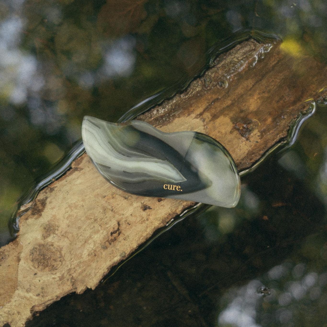 A translucent teardrop-shaped cosmetic tube or dropper cap with a marbled black, gray, and white pattern, featuring the brand name 'cure.' in gold text, resting on a wet log in shallow water.
