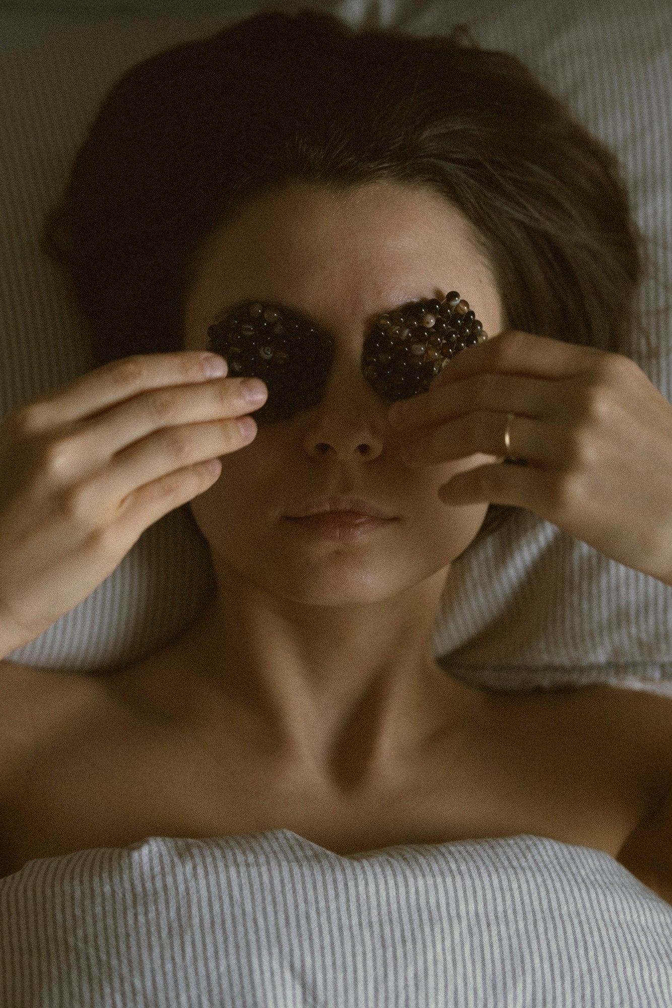 A woman lying in bed with her eyes covered by two dark, agate stone compresses, holding them in place with both hands. She is wearing a gold ring on one finger, and the background consists of soft, light-colored bedding.