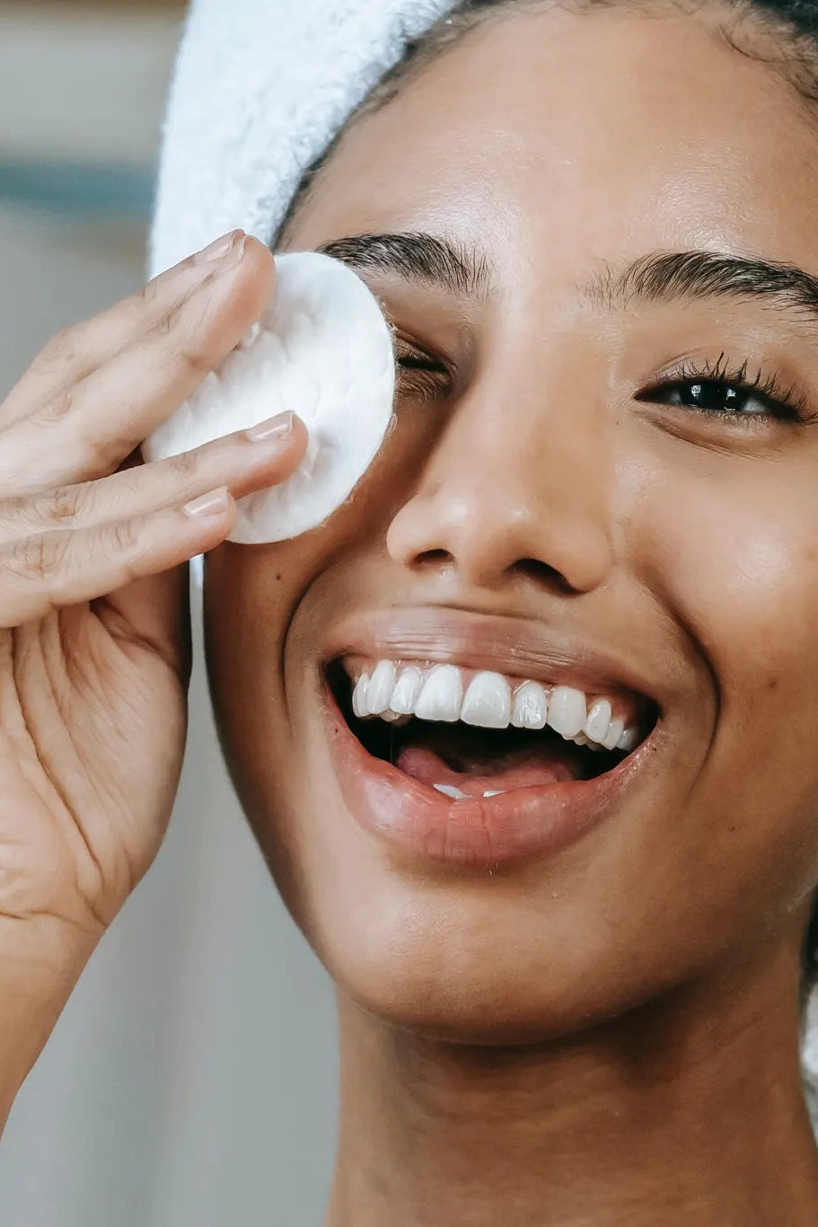 Woman applying cream to her face with a towel on her head, smiling.
