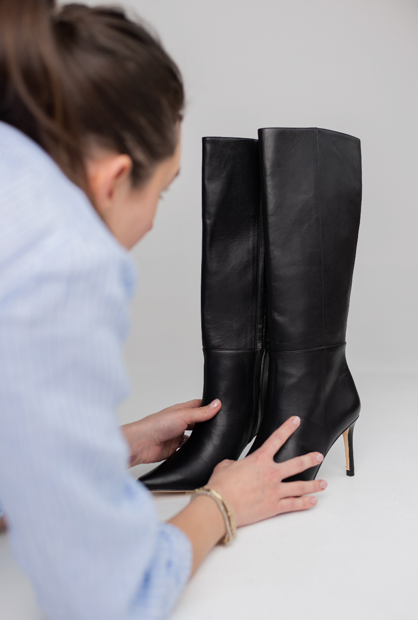 A person in a light blue shirt examines a pair of black leather knee-high boots with pointed toes and slim gold-tone heels against a clean light gray background.
