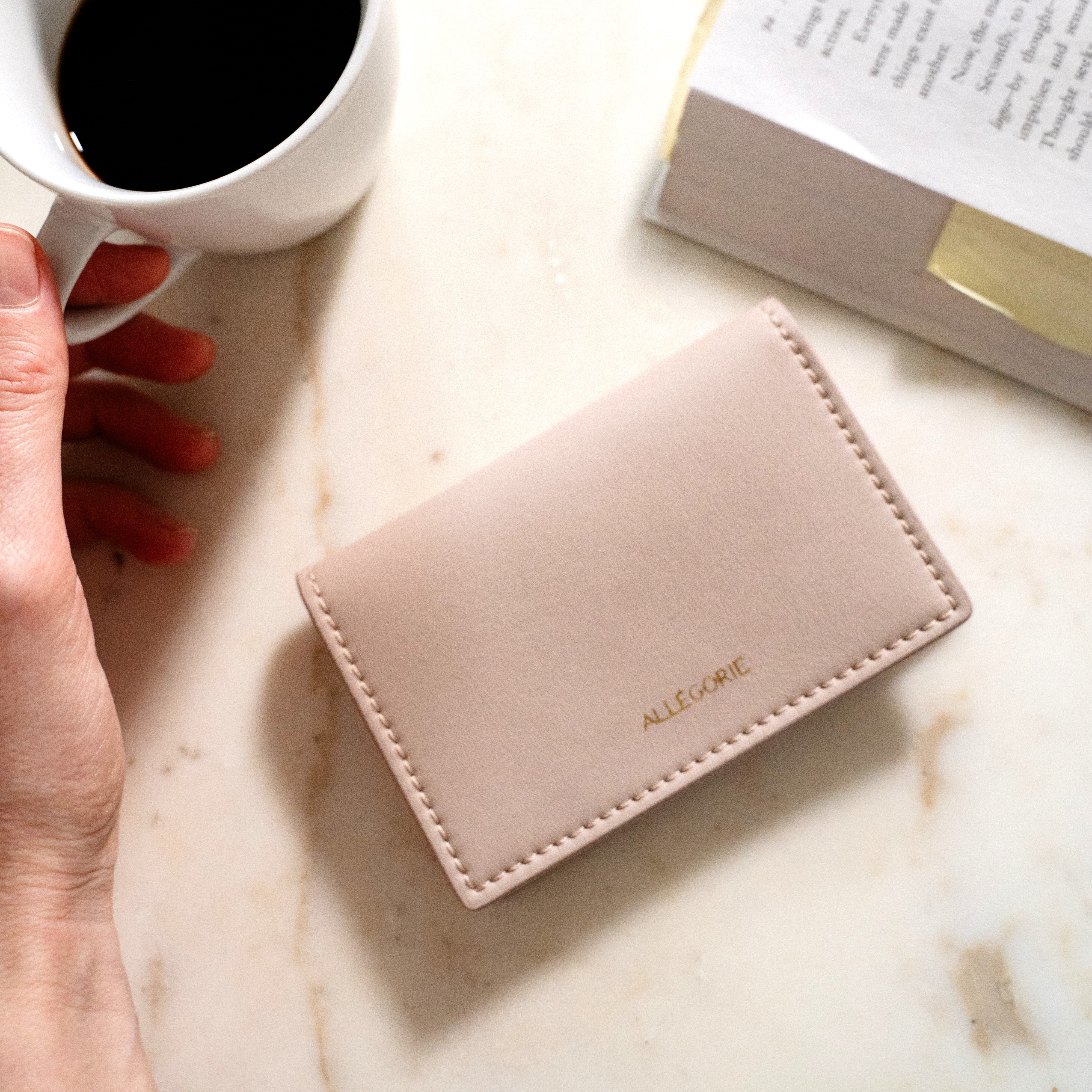 Pink cardholder with 'Allsop' branding on a marble surface next to a cup of coffee and an open book.