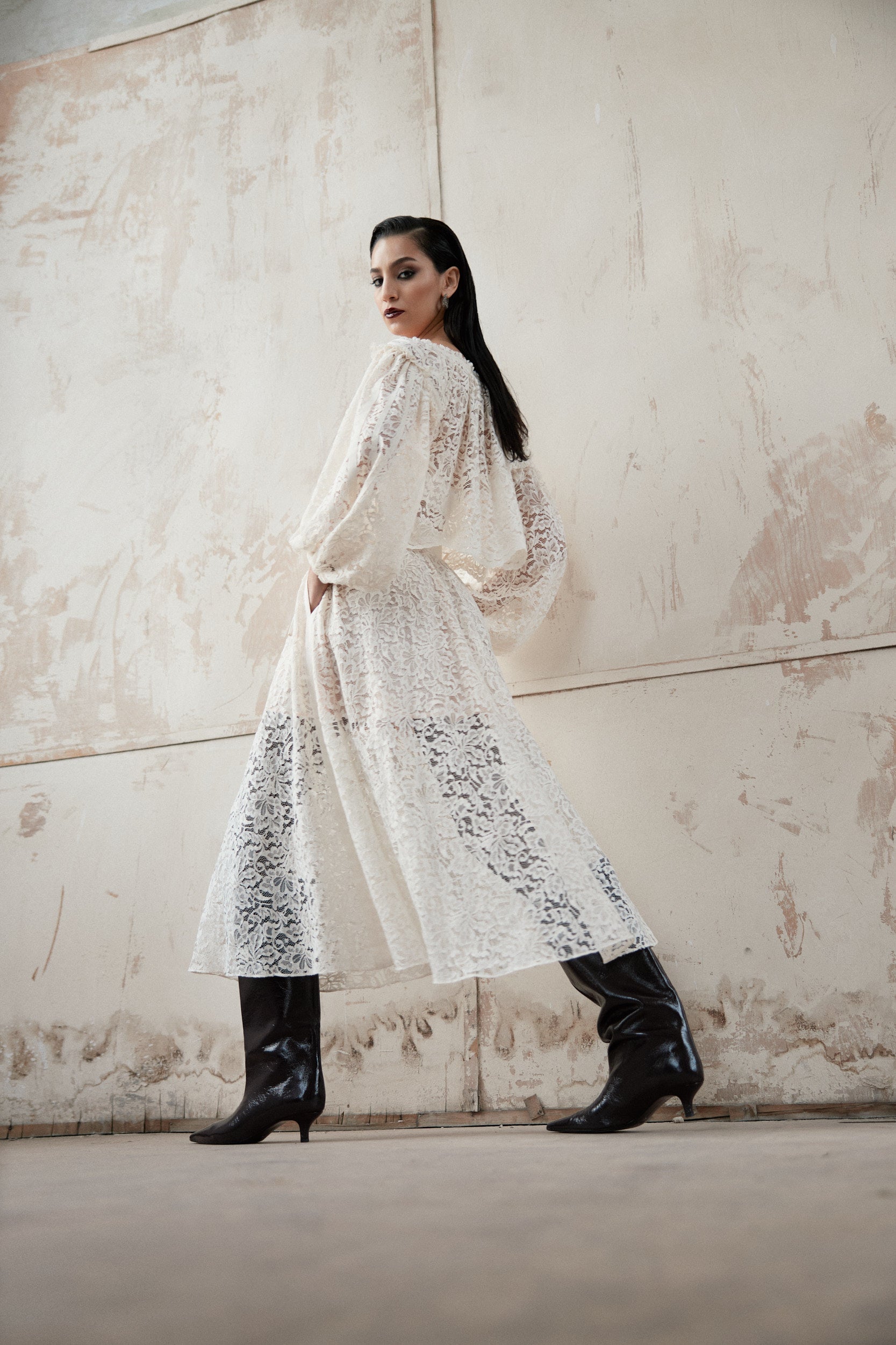 Woman in a long white lace dress and black boots standing against a textured wall.