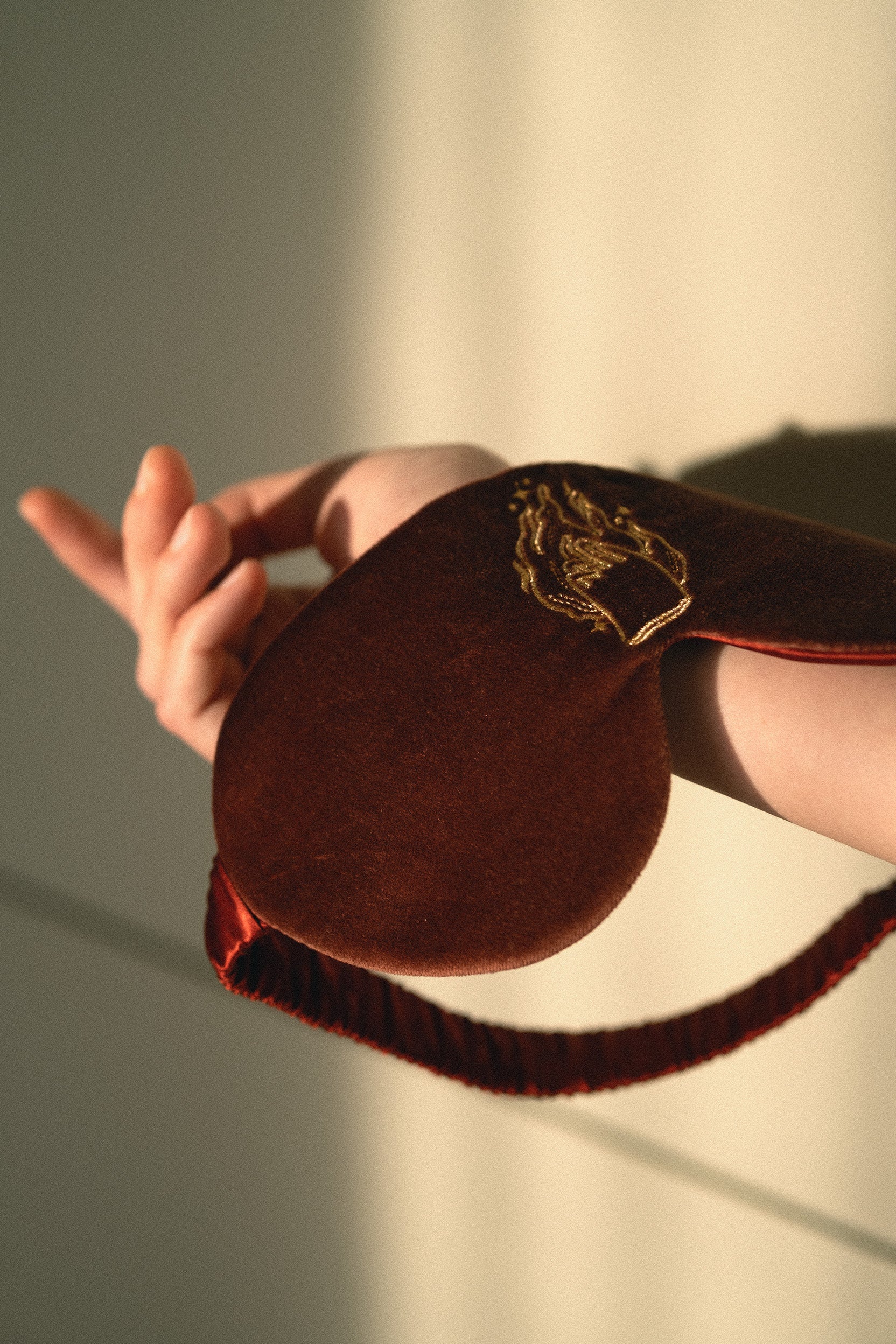 A close-up of a person's hand holding a dark brown velvet sleep mask with gold embroidered detailing and a matching dark red elastic strap, set against a warm-toned beige background with soft shadows.