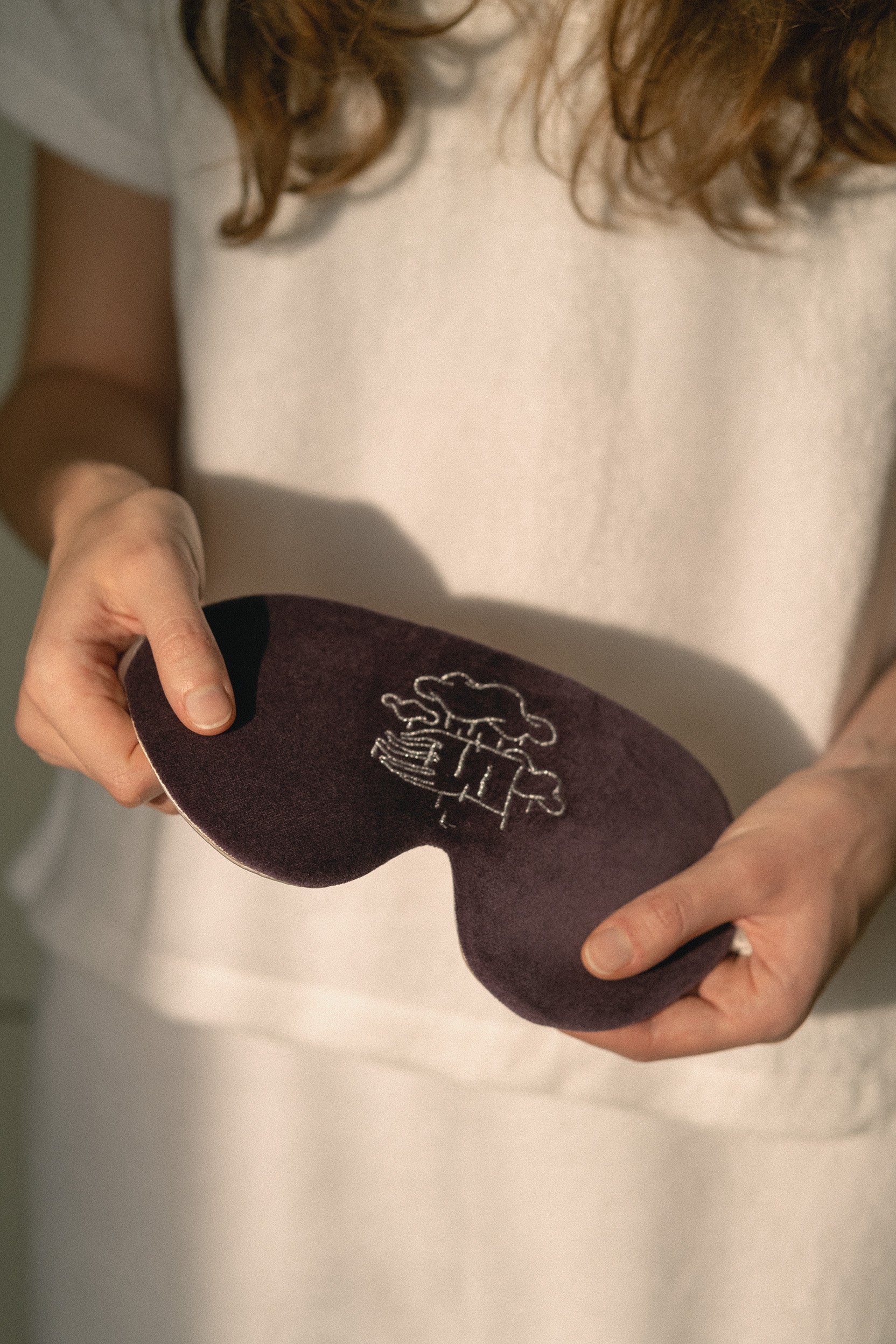 A person in a white shirt holds a dark plum-colored sleep mask with a white embroidered illustration of a bed and tree. The background is a soft, out-of-focus white.
