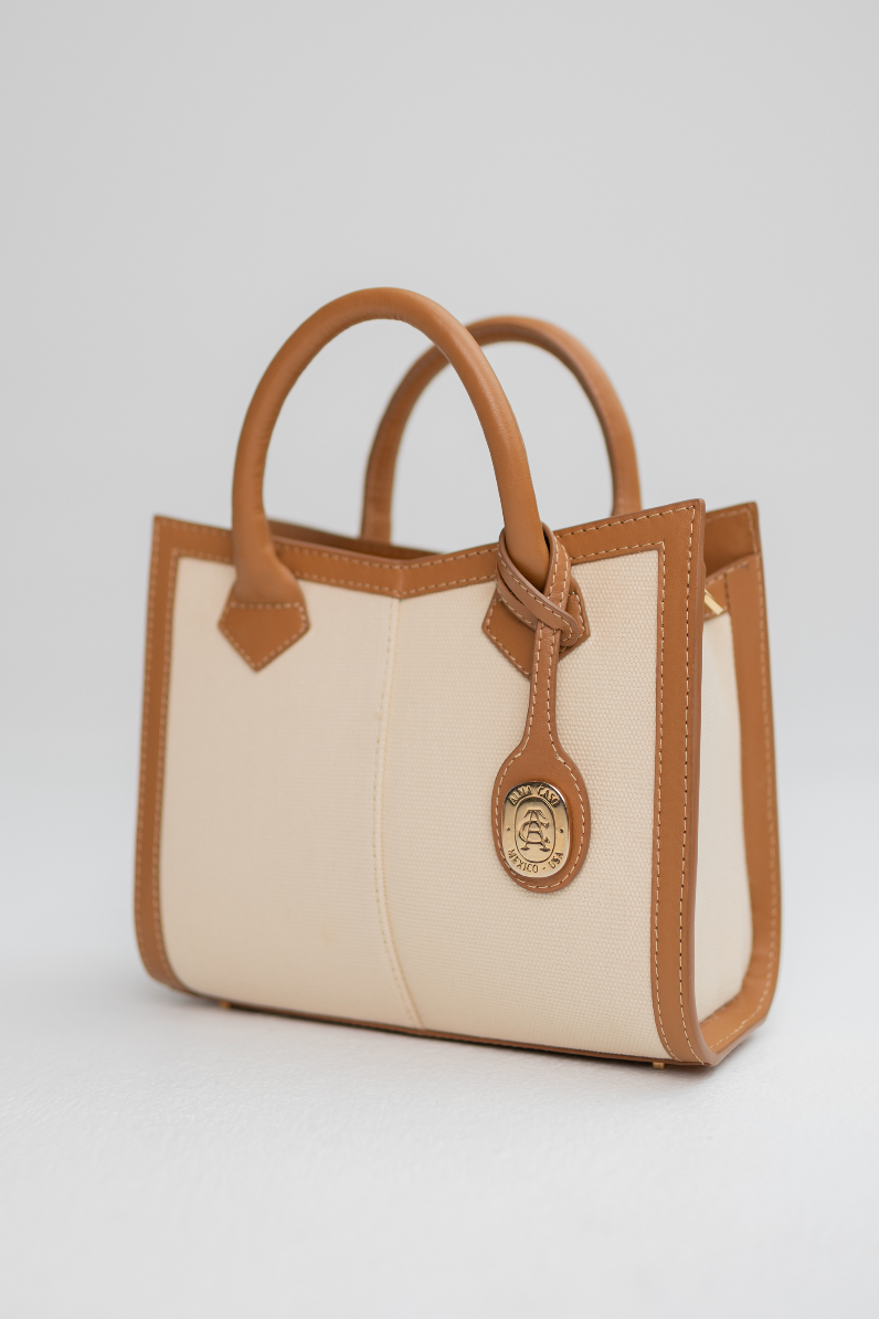 A structured mini tote bag with cream-colored panels and tan leather trim, featuring dual top handles, a decorative leather tag, and a round gold-tone logo medallion on the front, set against a neutral light gray background.