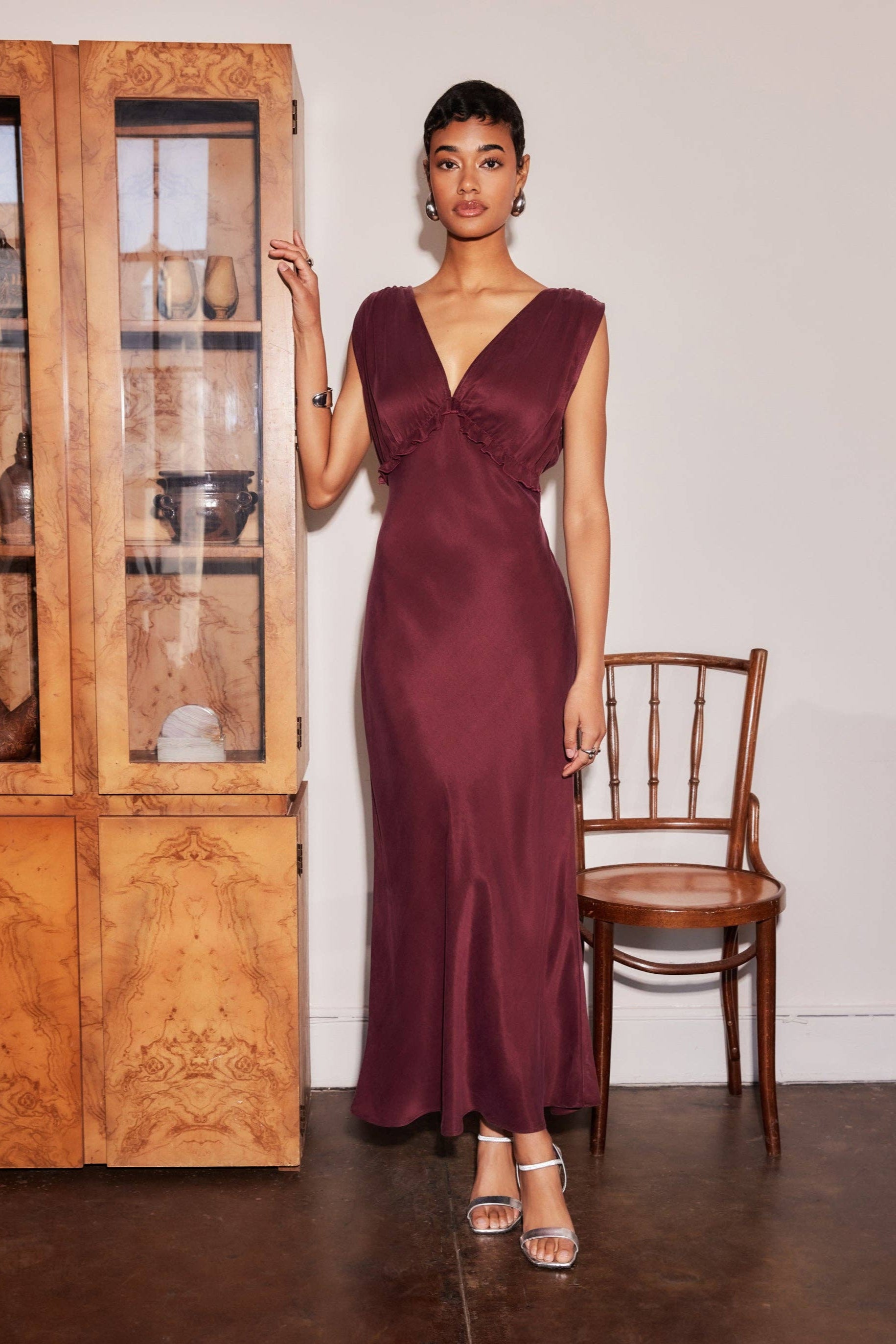 Woman in a burgundy evening gown standing next to a wooden chair and cabinet.