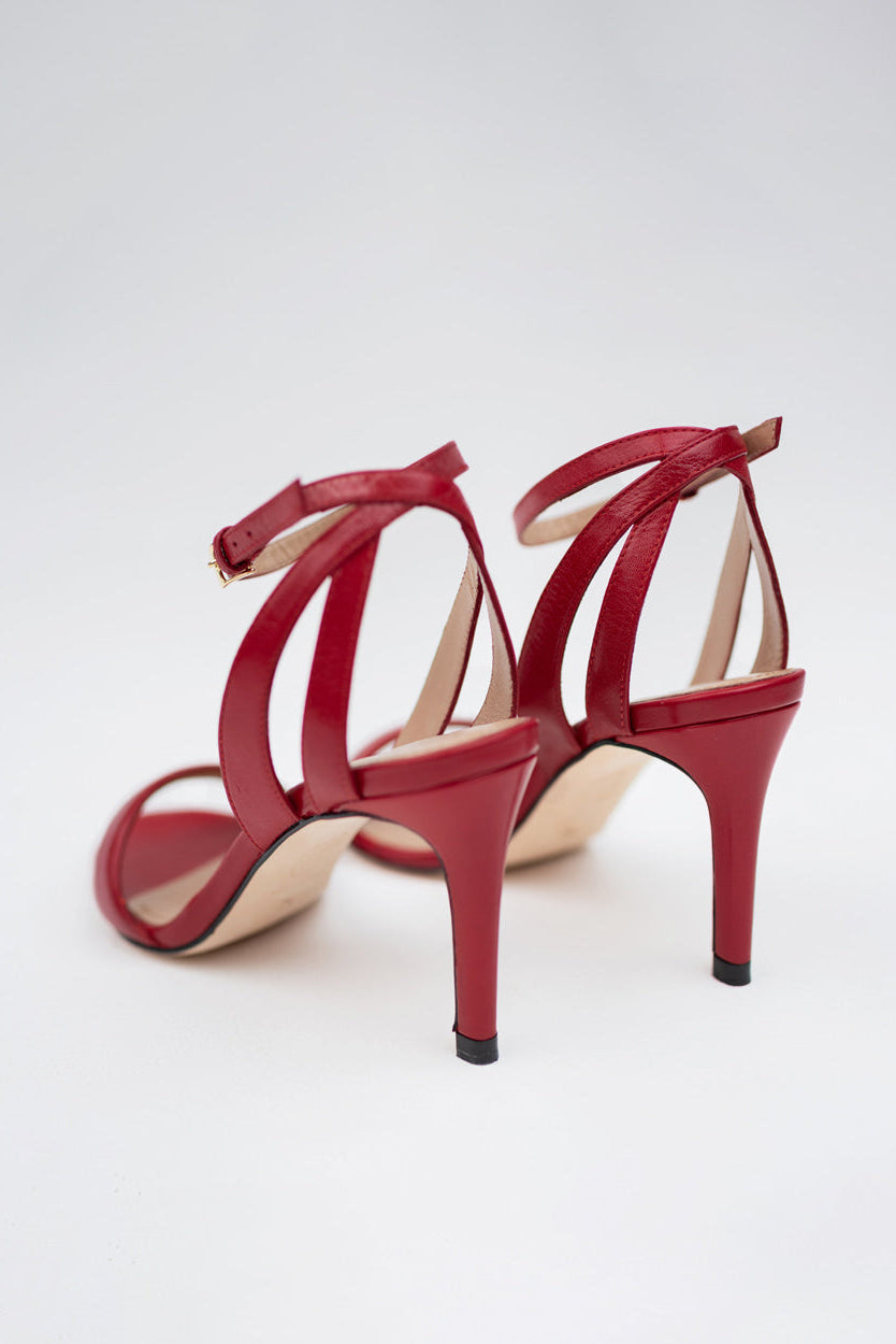 A pair of red strappy high-heeled sandals with ankle straps and stiletto heels, photographed from the back on a light gray studio background.